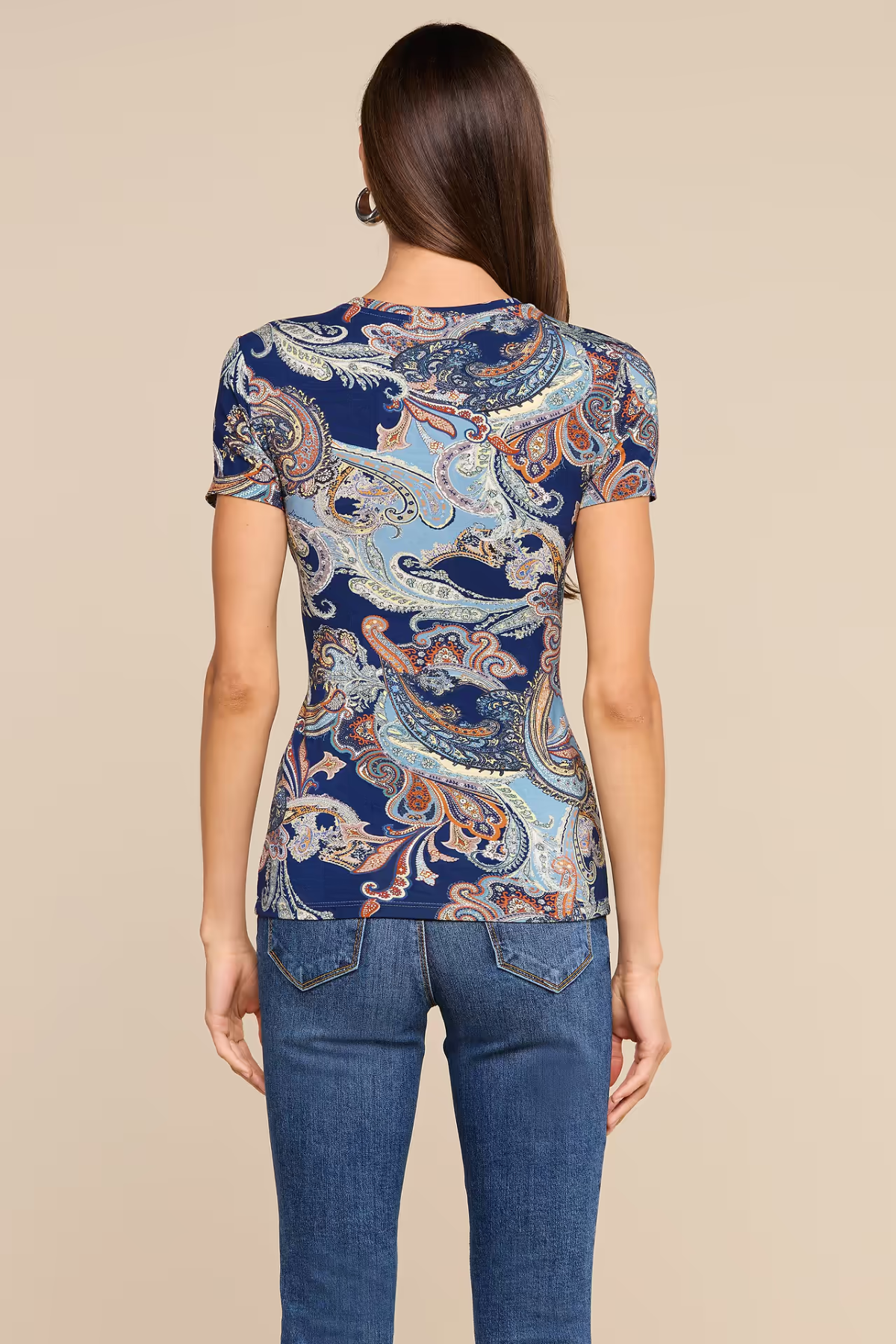 A woman viewed from the back wearing a fitted short-sleeve top with an ornate paisley-style print in navy, light blue, beige, and rust tones, paired with blue jeans against a plain beige studio background.