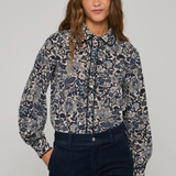 A woman modeling a long-sleeve button-up shirt with a navy blue and gray botanical pattern, paired with dark navy jeans, against a solid light gray background.