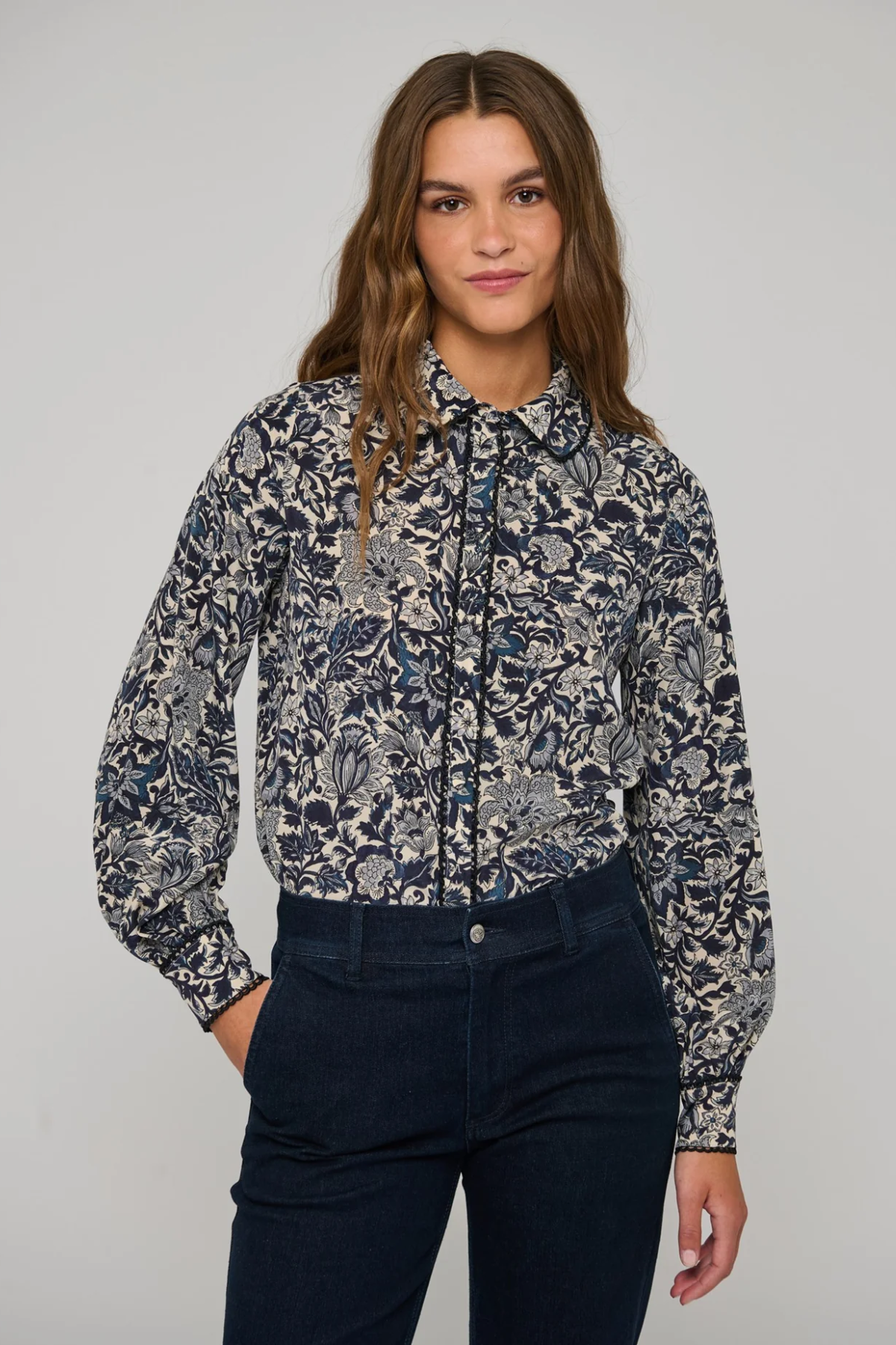 A woman modeling a long-sleeve button-up shirt with a navy blue and gray botanical pattern, paired with dark navy jeans, against a solid light gray background.