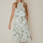 A woman modeling a sleeveless midi dress with vertical white and green stripes and a green floral print, featuring a matching tie belt and brown strappy sandals, set against a solid beige background.
