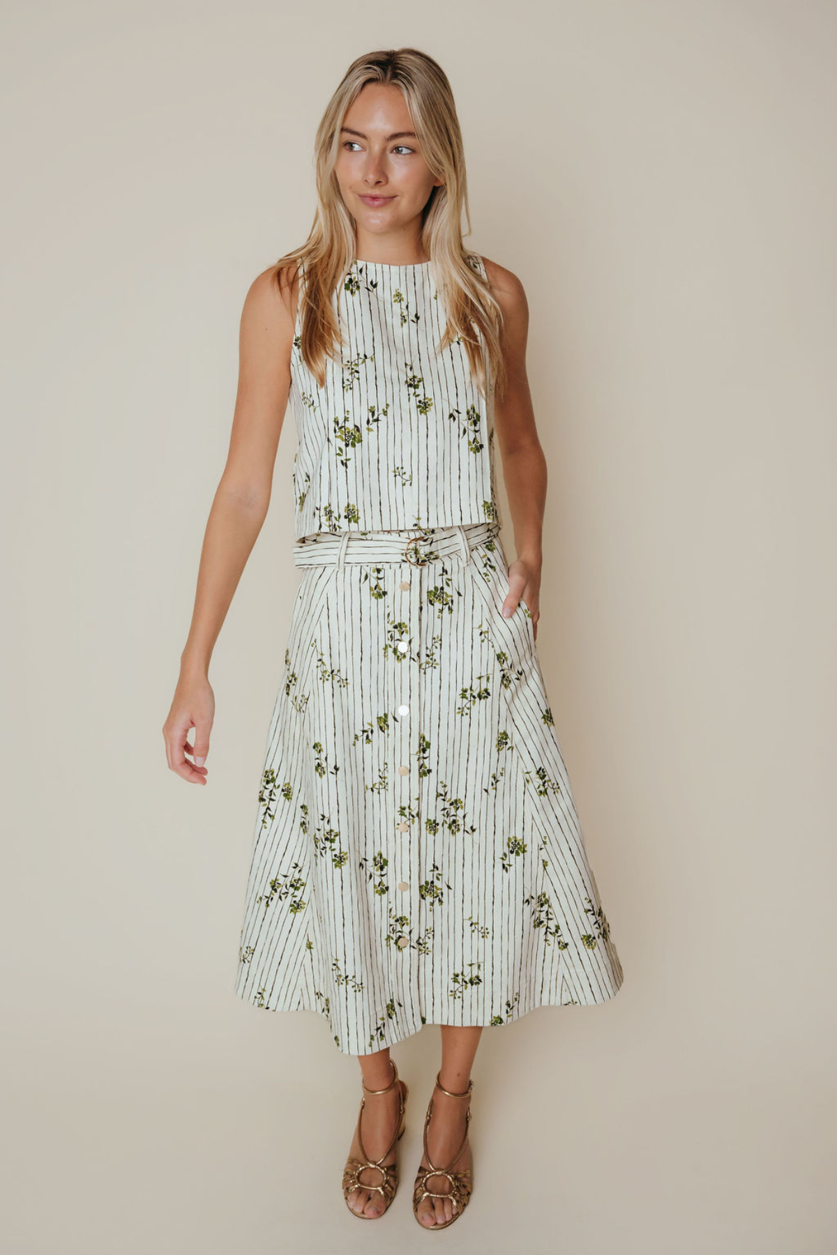 A woman modeling a sleeveless midi dress with vertical white and green stripes and a green floral print, featuring a matching tie belt and brown strappy sandals, set against a solid beige background.