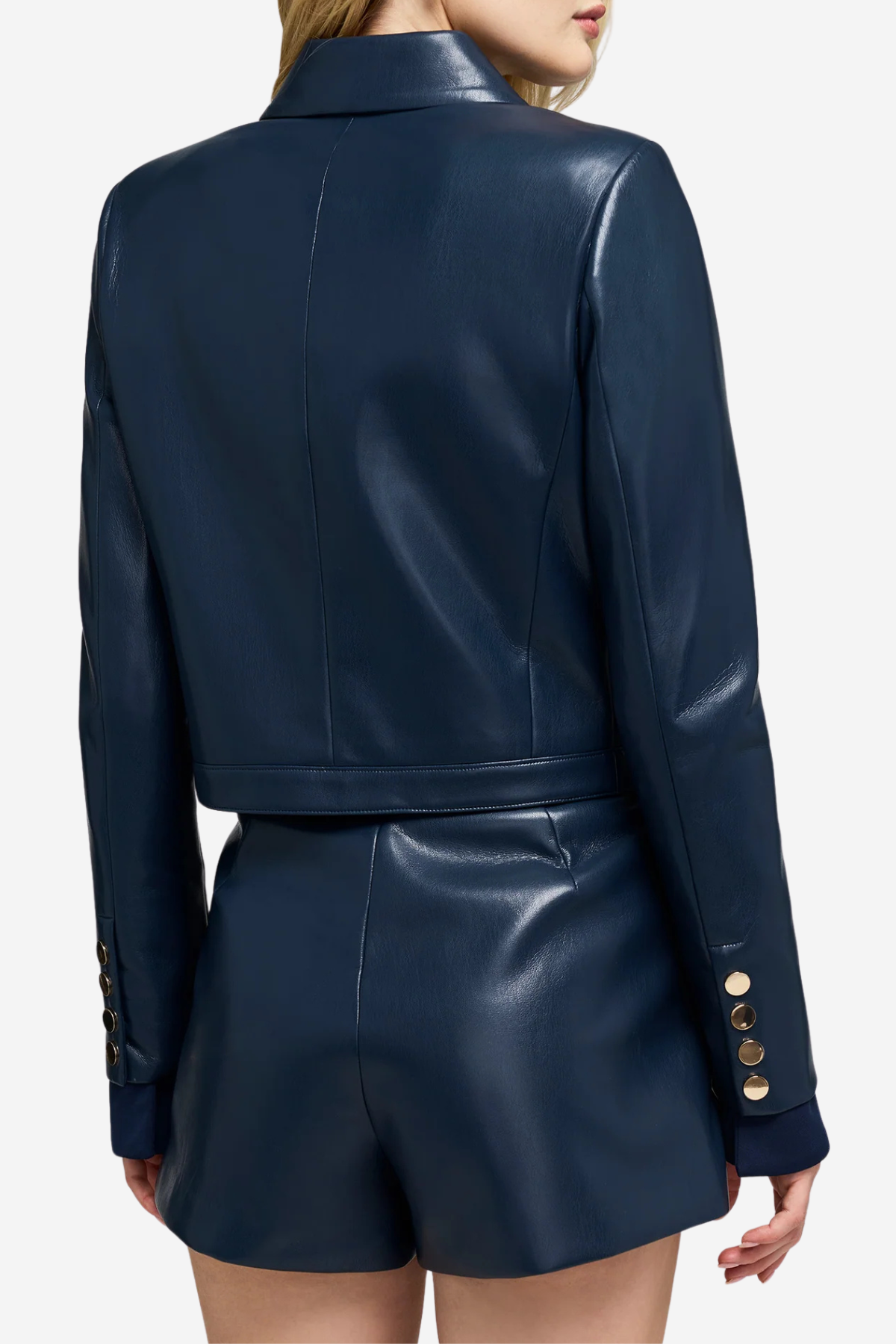 A model shown from the back wearing a dark navy faux-leather cropped jacket and matching shorts, featuring gold button details on the cuffs.
