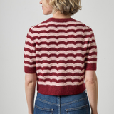 A woman shown from the back wearing a short-sleeve knit sweater with horizontal wavy stripes in burgundy and cream, paired with blue jeans, against a light gray studio background.