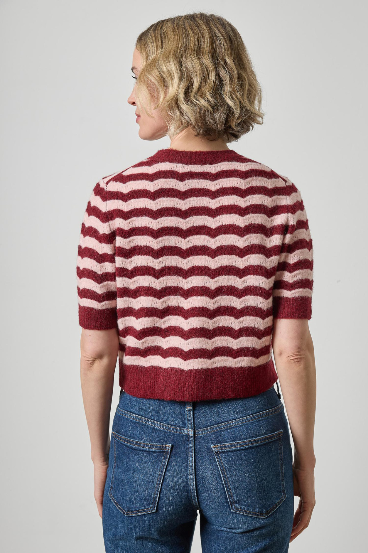 A woman shown from the back wearing a short-sleeve knit sweater with horizontal wavy stripes in burgundy and cream, paired with blue jeans, against a light gray studio background.