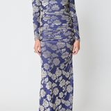 A woman modeling a long-sleeve, floor-length ruched dress with a blue and silver floral jacquard pattern, paired with black strappy sandals against a light gray studio background.