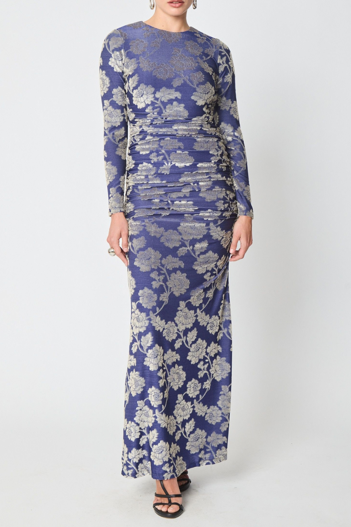 A woman modeling a long-sleeve, floor-length ruched dress with a blue and silver floral jacquard pattern, paired with black strappy sandals against a light gray studio background.
