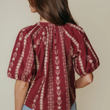 A woman shown from the back wearing a burgundy puff-sleeve blouse with vertical cream floral patterns, paired with light blue jeans, against a solid beige background.
