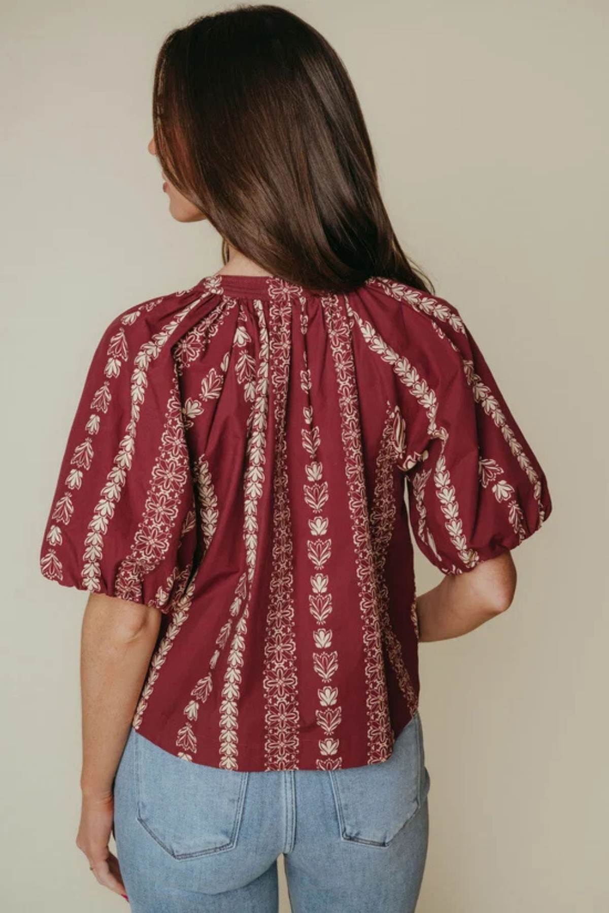 A woman shown from the back wearing a burgundy puff-sleeve blouse with vertical cream floral patterns, paired with light blue jeans, against a solid beige background.