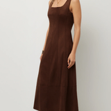 A woman modeling a sleeveless brown midi dress with a square neckline and side pockets, paired with snakeskin-patterned boots, against a plain light gray studio background.