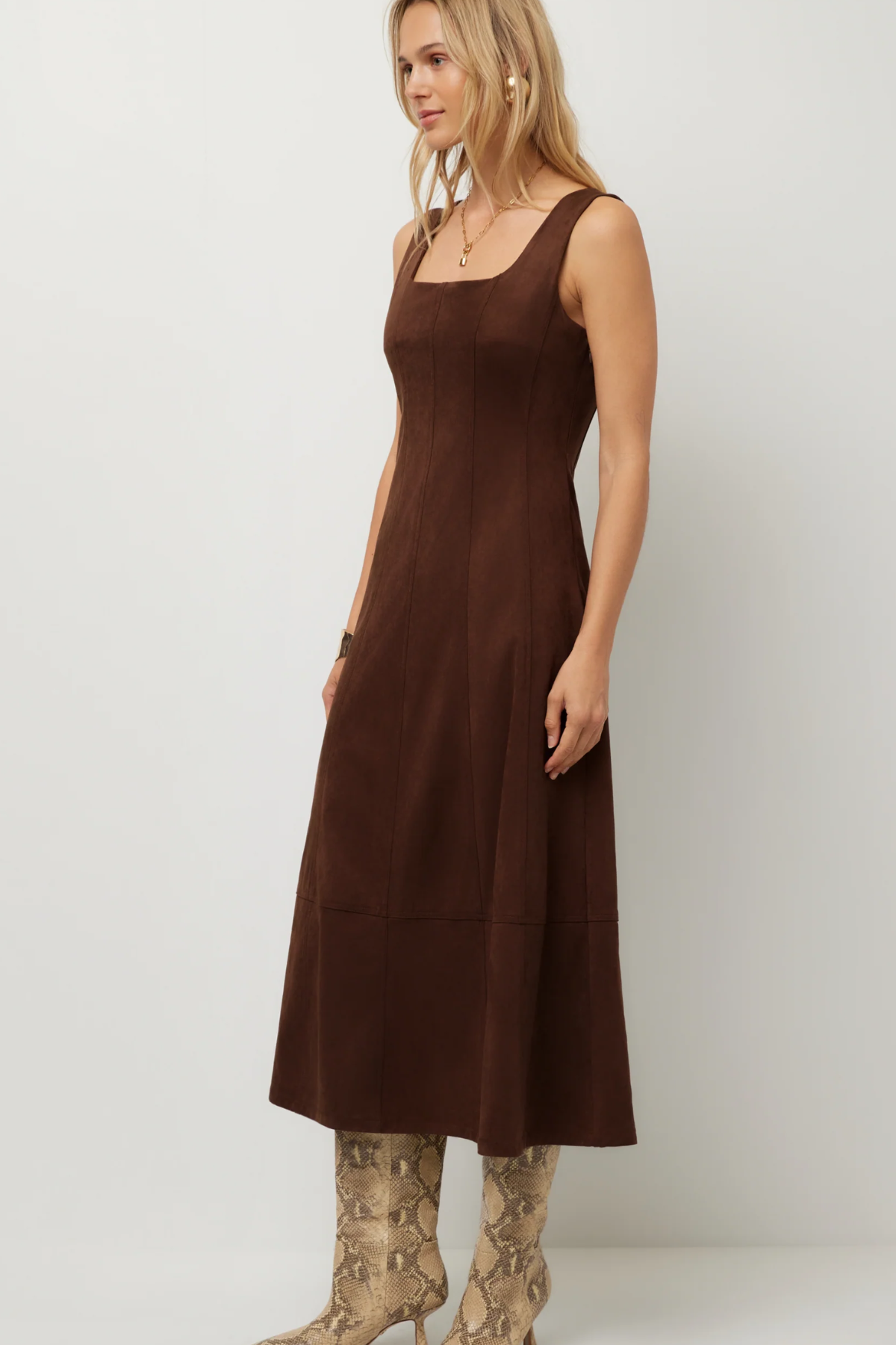 A woman modeling a sleeveless brown midi dress with a square neckline and side pockets, paired with snakeskin-patterned boots, against a plain light gray studio background.