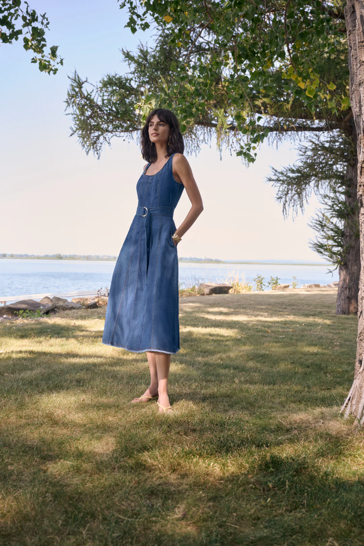 Woman in a blue dress standing in a grassy area near a body of water with trees in the background.