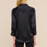 A woman viewed from the back wearing a black satin button-up blouse and black pants against a plain beige studio background.