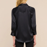 A woman viewed from the back wearing a black satin button-up blouse and black pants against a plain beige studio background.