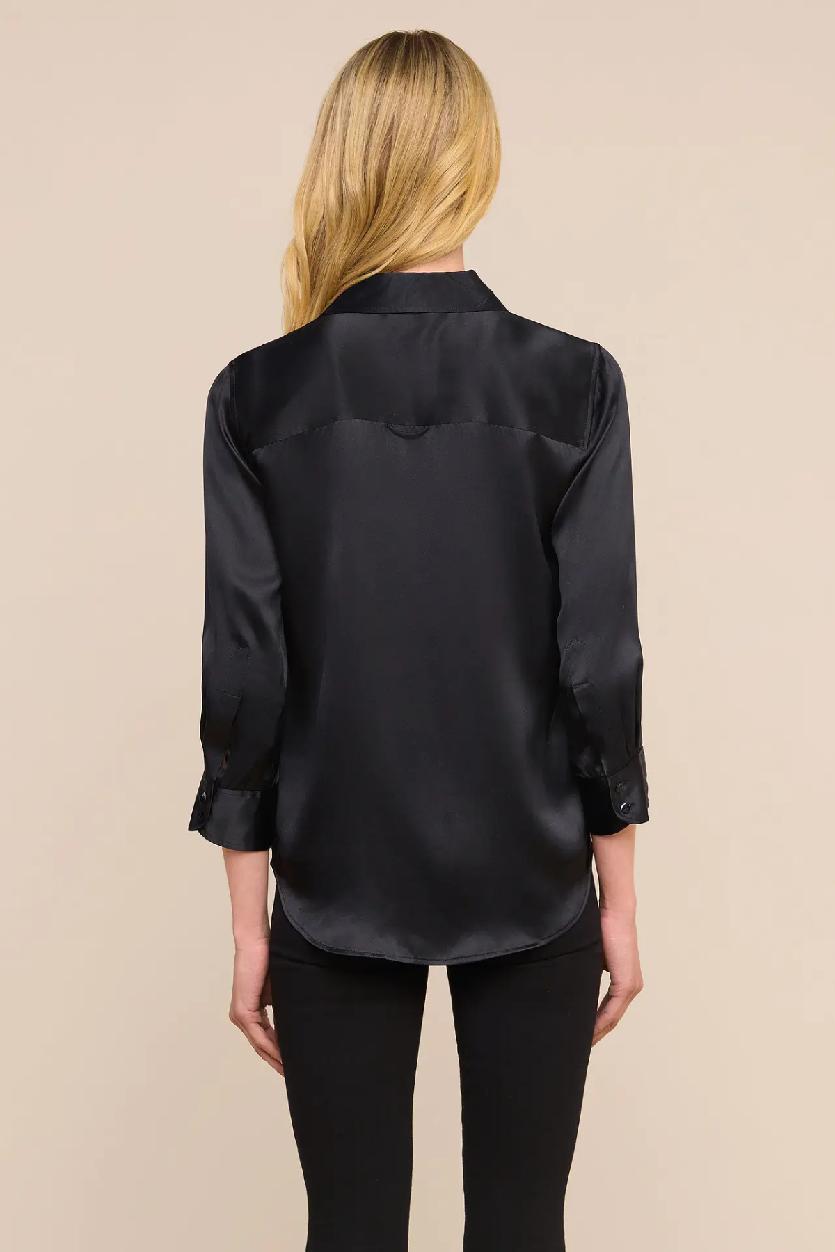 A woman viewed from the back wearing a black satin button-up blouse and black pants against a plain beige studio background.