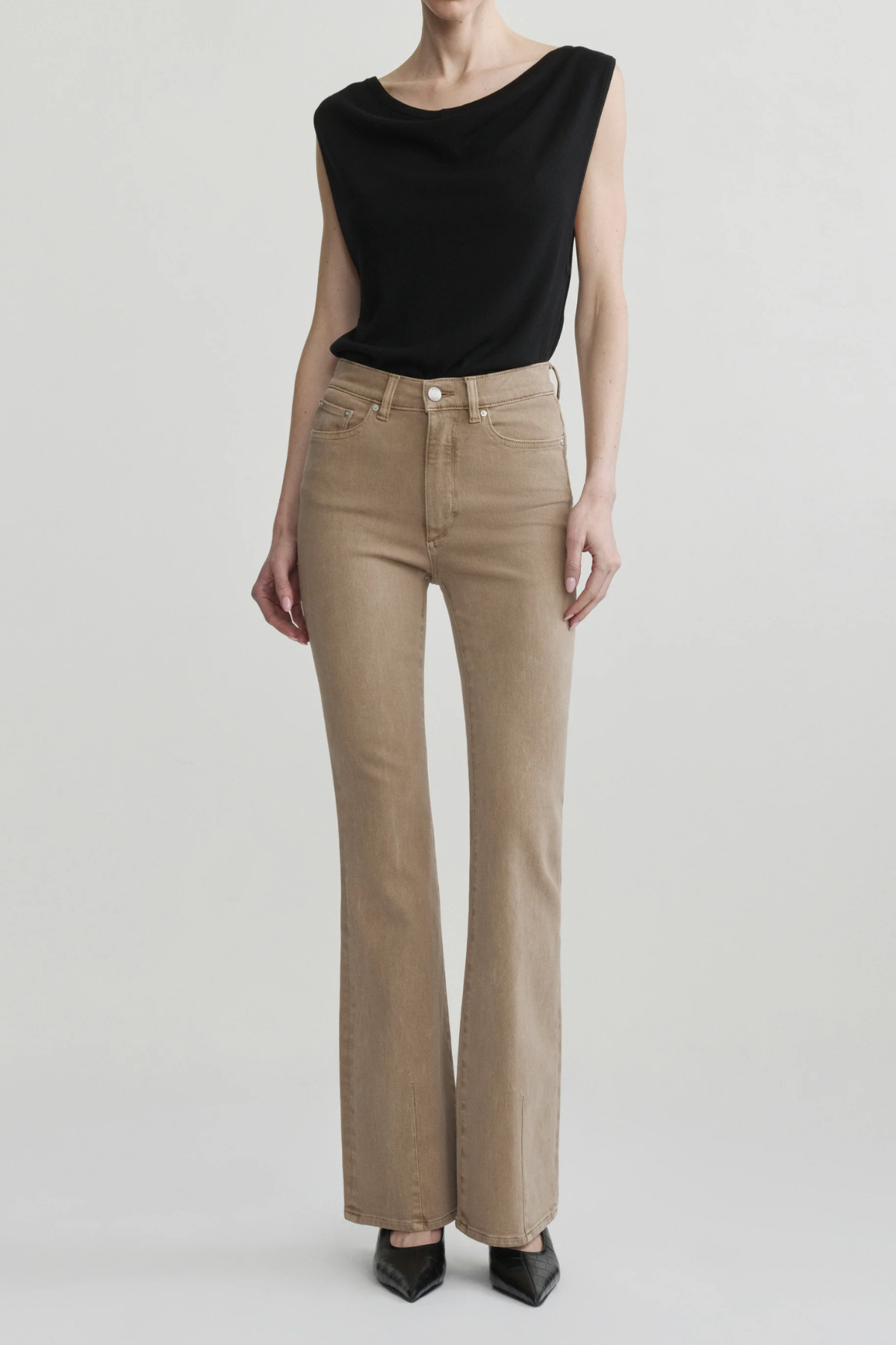 A model wearing beige flared jeans and a black sleeveless top, standing against a light gray studio background.