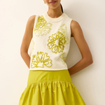Woman wearing a white sleeveless top with floral patterns and a yellow skirt against a plain background