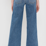Back view of a person wearing blue wide-leg denim jeans and beige woven flat shoes against a plain white studio background.