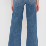 Back view of a person wearing blue wide-leg denim jeans and beige woven flat shoes against a plain white studio background.