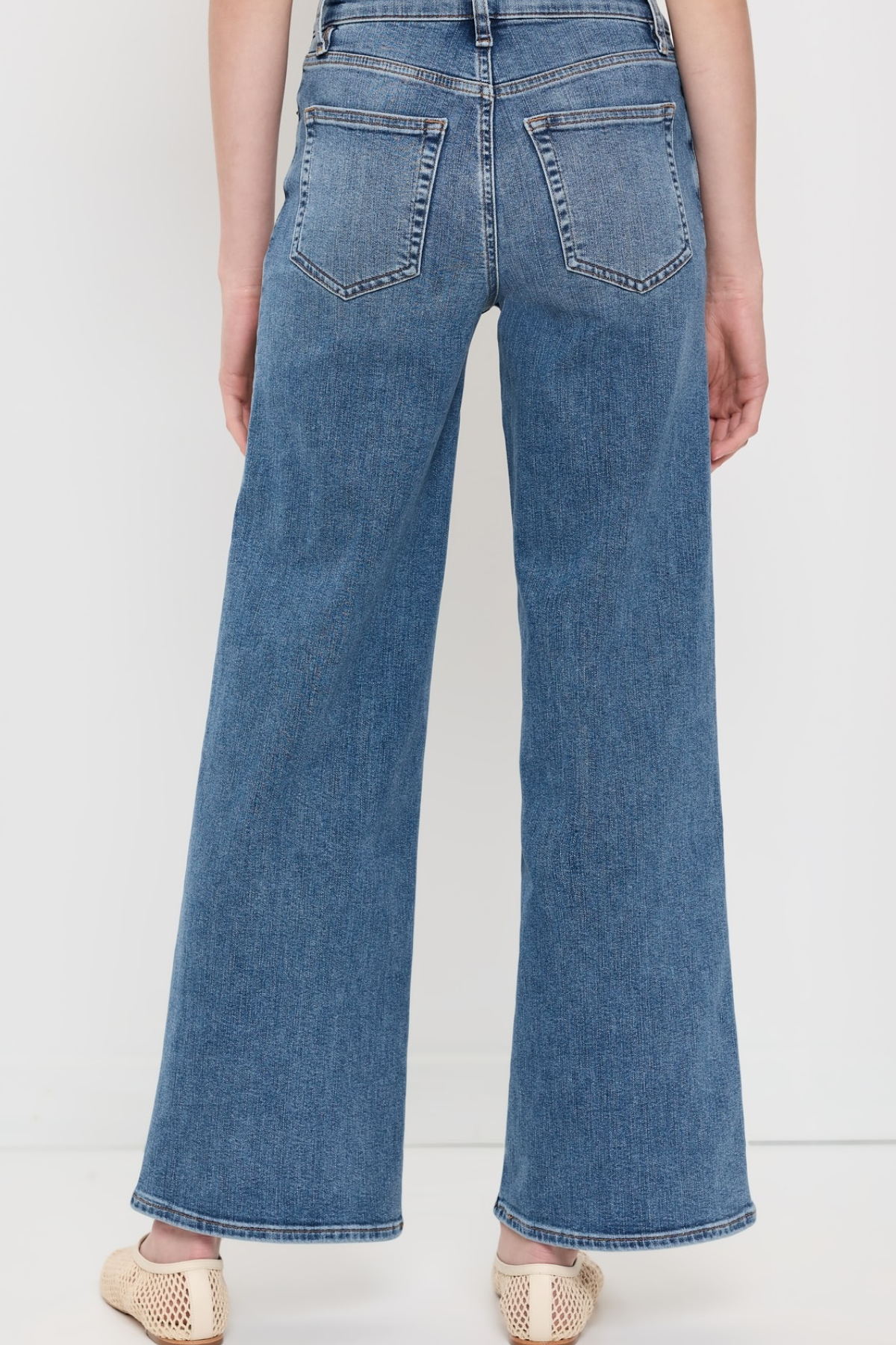 Back view of a person wearing blue wide-leg denim jeans and beige woven flat shoes against a plain white studio background.