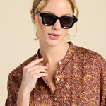 A woman wearing black cat-eye sunglasses and a brown floral button-up shirt, photographed against a solid beige background.