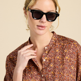 A woman wearing black cat-eye sunglasses and a brown floral button-up shirt, photographed against a solid beige background.