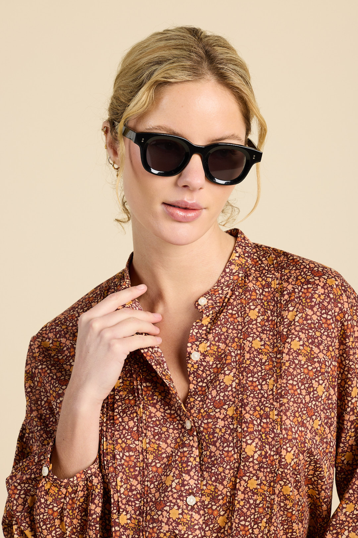 A woman wearing black cat-eye sunglasses and a brown floral button-up shirt, photographed against a solid beige background.