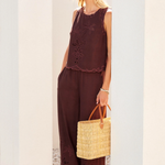 Woman in brown sleeveless top and wide-leg pants holding a straw bag against a white wall.
