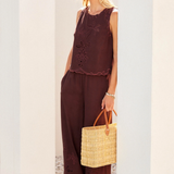 Woman in brown sleeveless top and wide-leg pants holding a straw bag against a white wall.