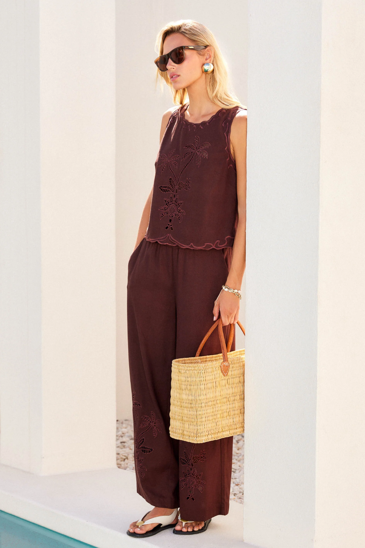 Woman in brown sleeveless top and wide-leg pants holding a straw bag against a white wall.