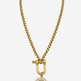 A gold-toned chain necklace featuring a large toggle clasp and a circular medallion pendant with a sunburst or floral pattern, shown against a plain light gray background.