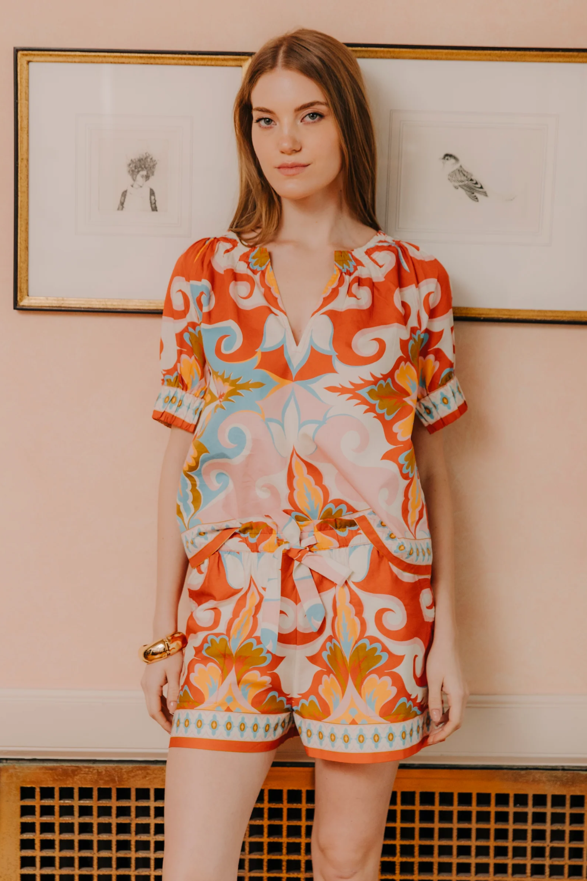 Woman wearing a colorful patterned outfit standing against a light pink wall with framed pictures.