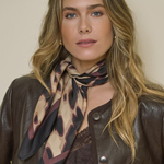 A woman with blonde hair wearing a brown leather jacket and a patterned scarf in beige, brown, and black, posed against a plain beige background.