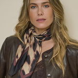 A woman with blonde hair wearing a brown leather jacket and a patterned scarf in beige, brown, and black, posed against a plain beige background.
