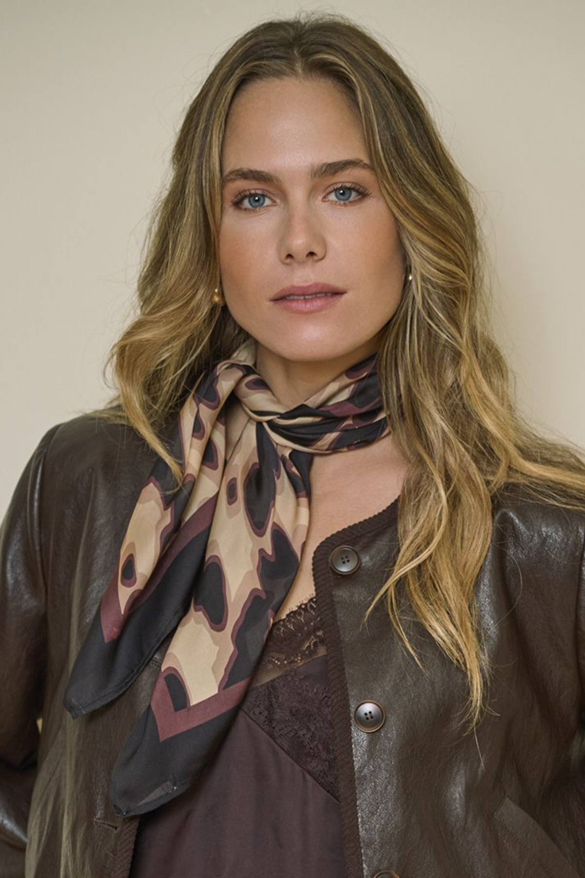 A woman with blonde hair wearing a brown leather jacket and a patterned scarf in beige, brown, and black, posed against a plain beige background.