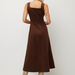 A woman shown from the back wearing a sleeveless brown midi dress with a square neckline and a tiered skirt, paired with snakeskin-patterned heeled boots, against a plain light gray studio background.