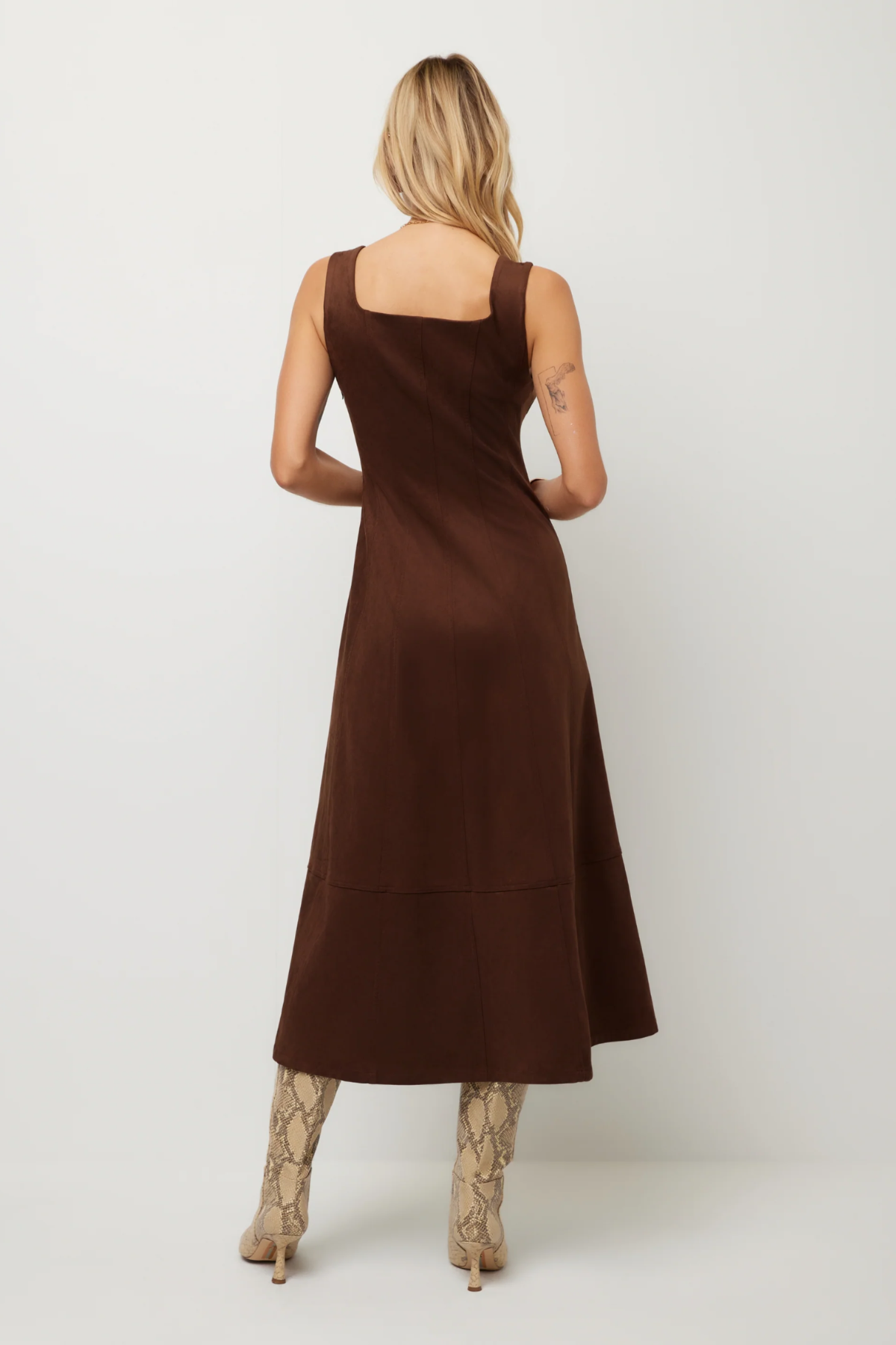 A woman shown from the back wearing a sleeveless brown midi dress with a square neckline and a tiered skirt, paired with snakeskin-patterned heeled boots, against a plain light gray studio background.