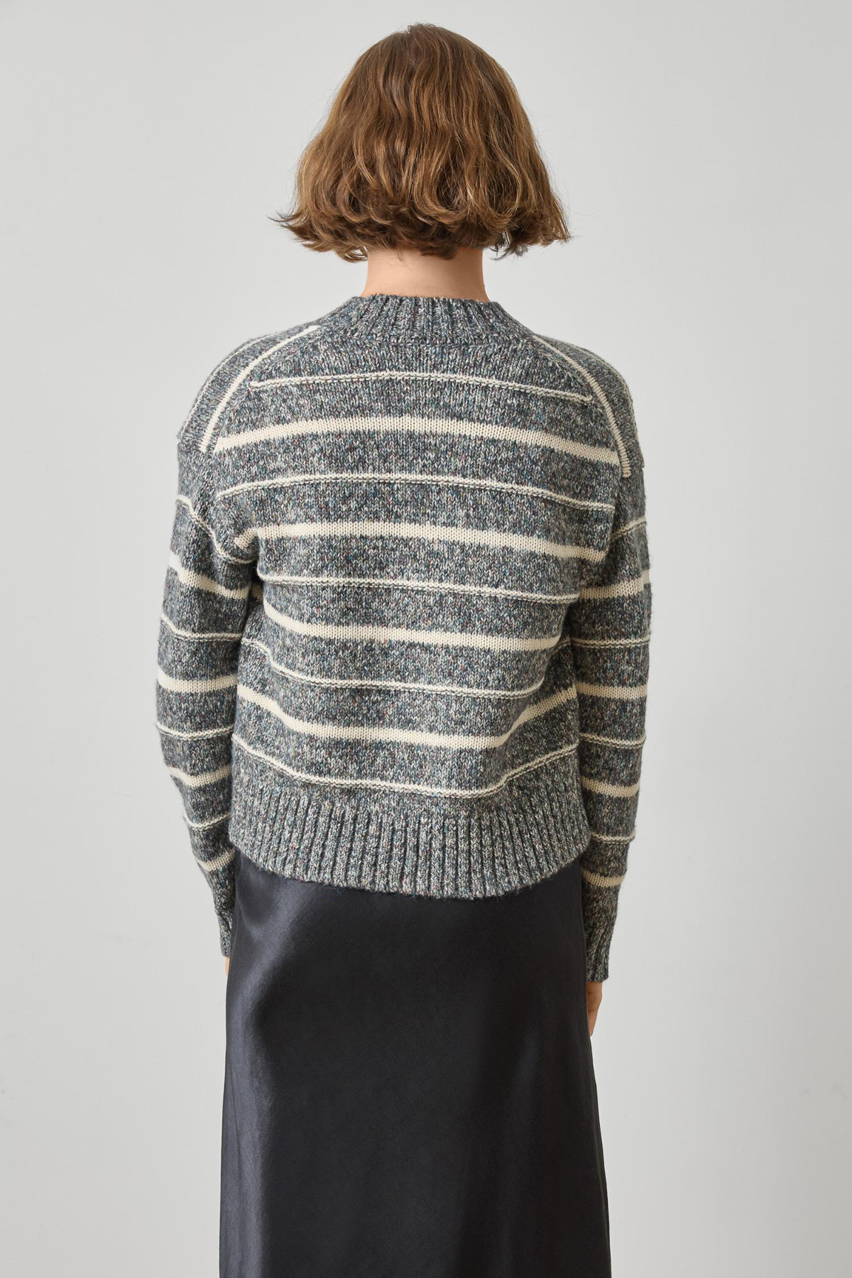 A model viewed from the back wearing a gray and cream striped knit sweater and a dark skirt, set against a plain light gray studio background.