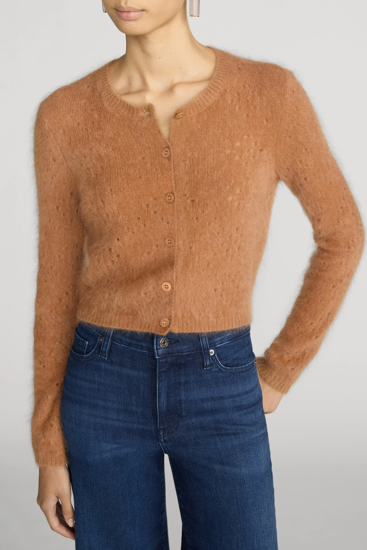 A woman wearing a fuzzy, cropped button-front cardigan in a warm camel brown color, paired with dark blue jeans, against a light gray studio background.
