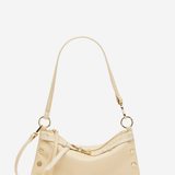 A light beige leather shoulder bag with gold-tone hardware, featuring a crescent-shaped body, a long shoulder strap, and decorative rivet details on the sides, set against a plain white background.