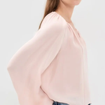 A woman wearing a light pink blouse with dramatic balloon sleeves and a gathered neckline, paired with blue jeans, photographed against a plain light gray studio background.