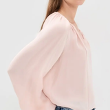A woman wearing a light pink blouse with dramatic balloon sleeves and a gathered neckline, paired with blue jeans, photographed against a plain light gray studio background.