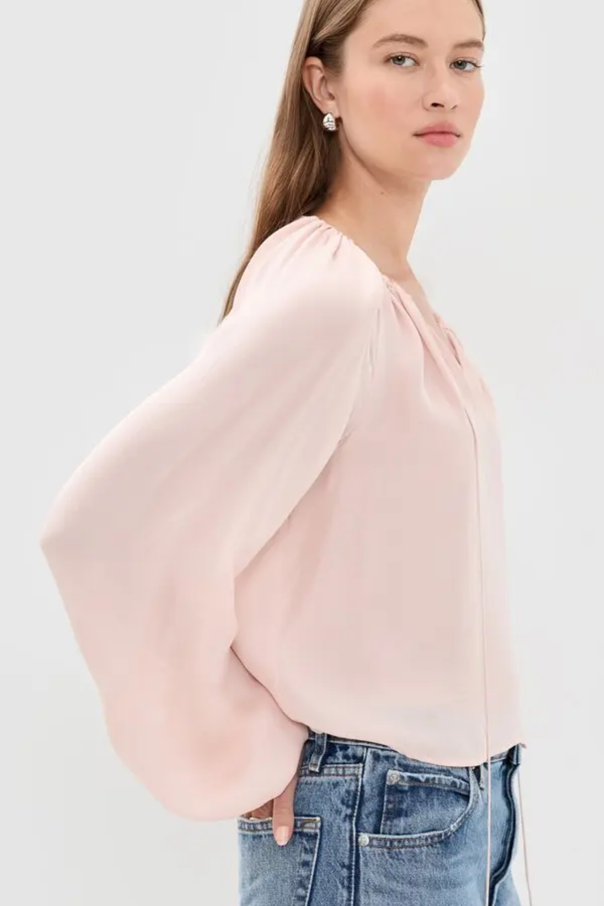 A woman wearing a light pink blouse with dramatic balloon sleeves and a gathered neckline, paired with blue jeans, photographed against a plain light gray studio background.