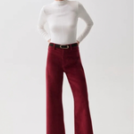 A woman modeling a white mock-neck long-sleeve top and burgundy wide-leg cropped trousers with a black belt, paired with black pointed-toe heels, against a light gray studio background.