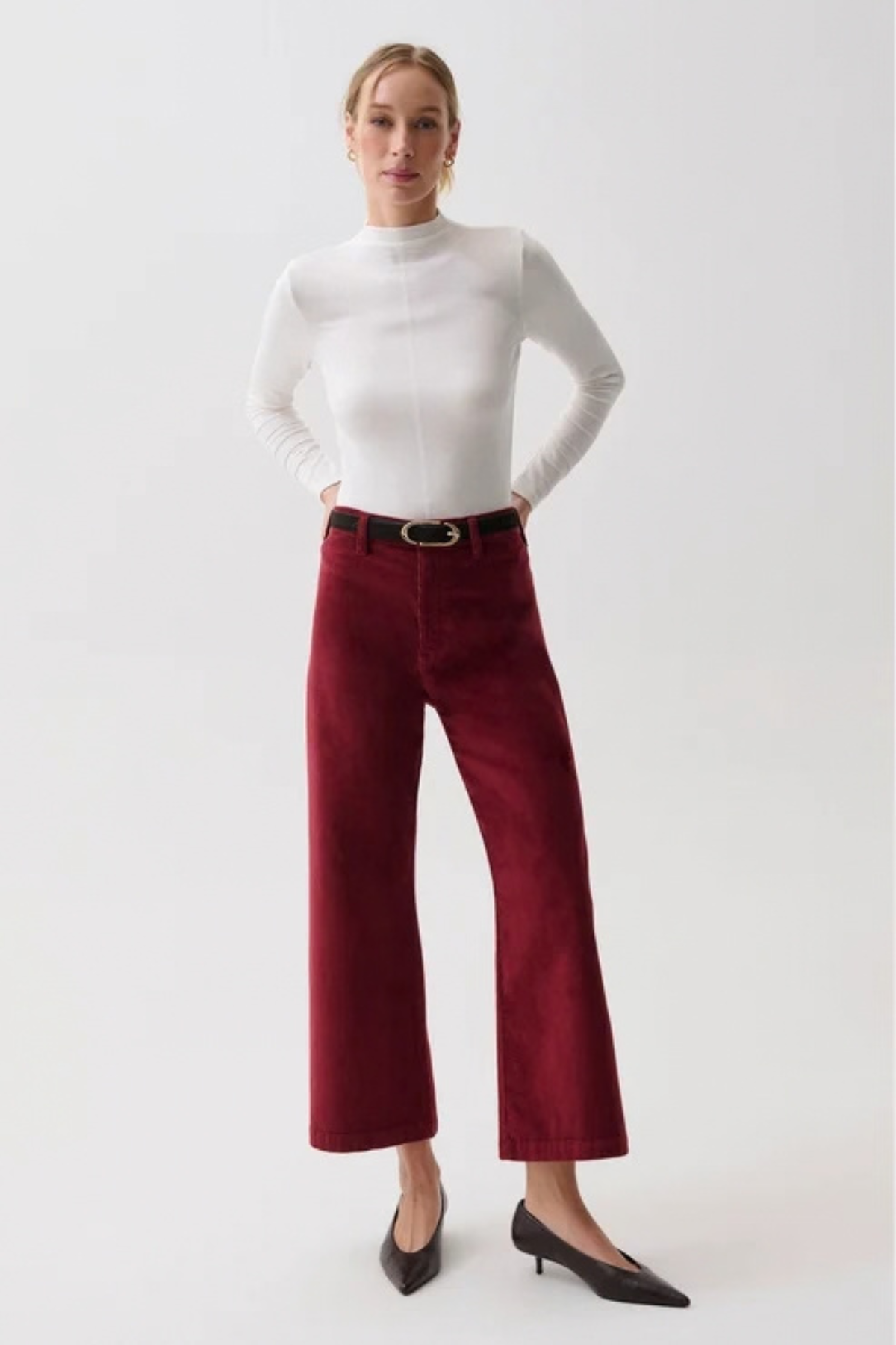 A woman modeling a white mock-neck long-sleeve top and burgundy wide-leg cropped trousers with a black belt, paired with black pointed-toe heels, against a light gray studio background.