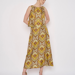 A woman modeling a sleeveless midi dress with a yellow, gold, and brown floral pattern, standing against a plain white studio background.