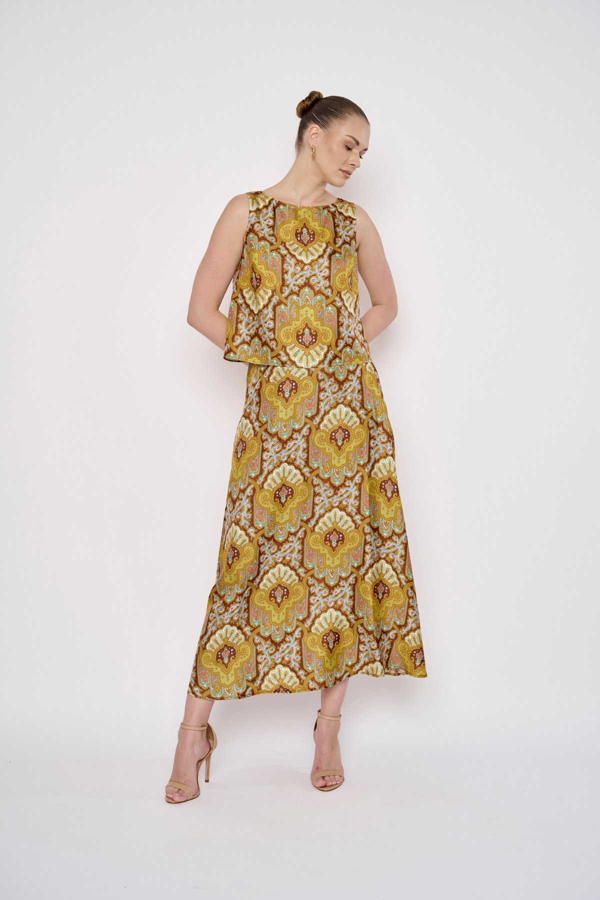 A woman modeling a sleeveless midi dress with a yellow, gold, and brown floral pattern, standing against a plain white studio background.
