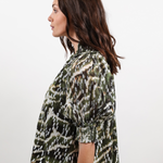 A woman shown in side profile wearing a short-sleeve blouse with an abstract camouflage-like pattern in olive green, black, and off-white, paired with blue jeans against a plain light gray studio background.