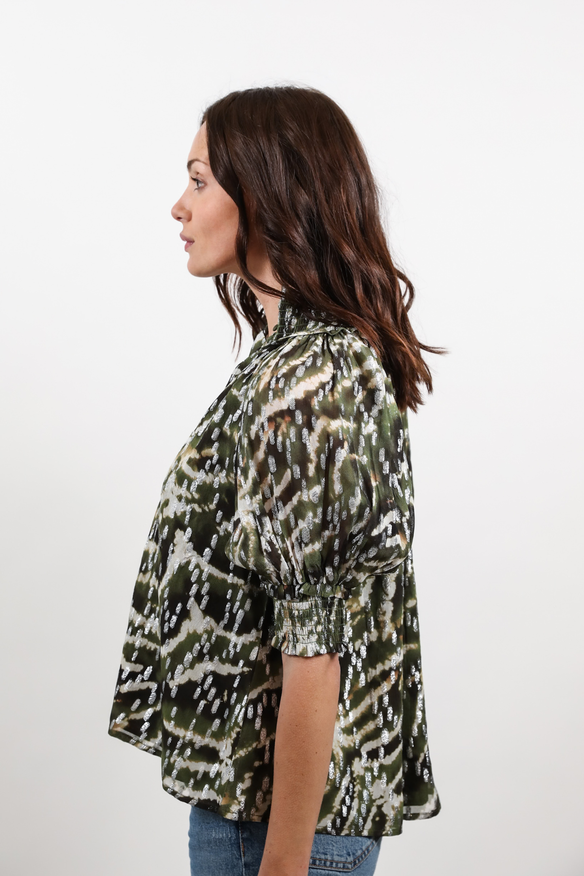 A woman shown in side profile wearing a short-sleeve blouse with an abstract camouflage-like pattern in olive green, black, and off-white, paired with blue jeans against a plain light gray studio background.