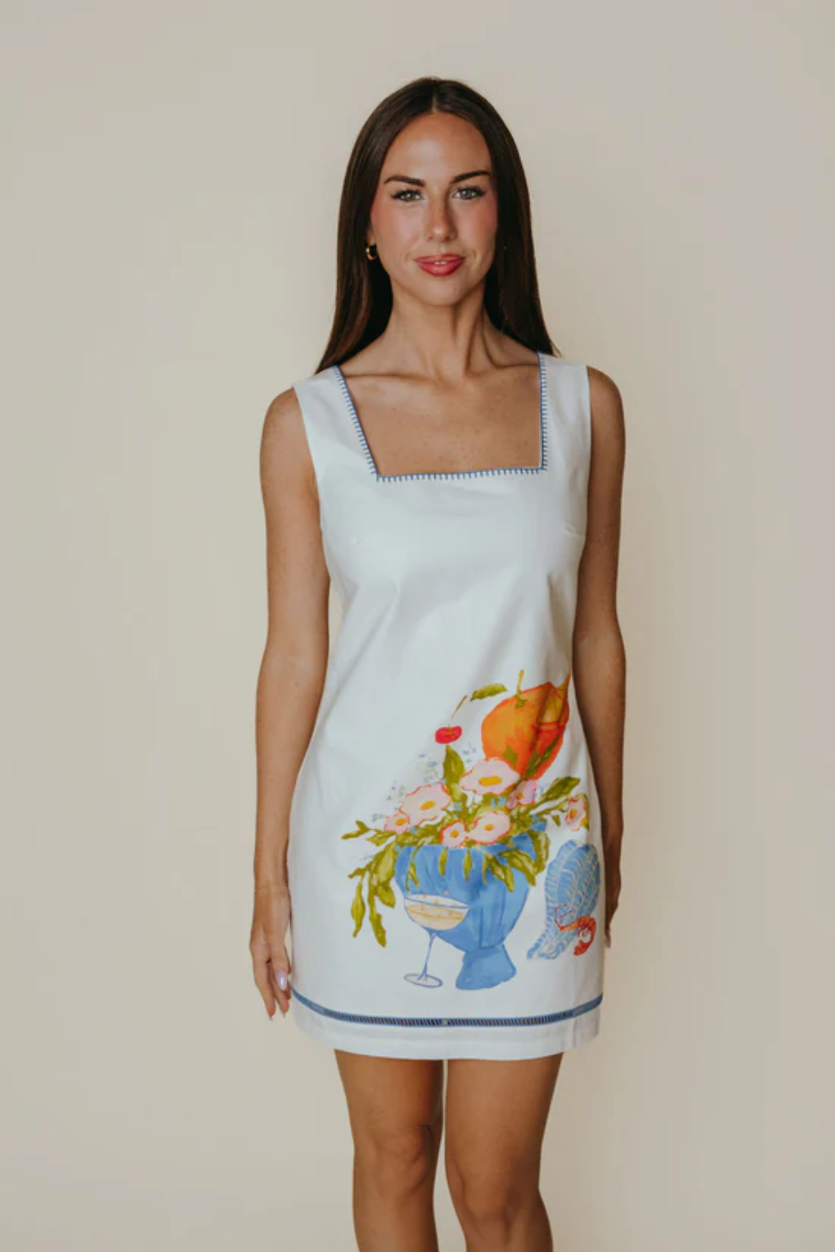 Woman wearing a sleeveless dress with floral and fruit design on a plain background