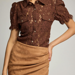 A model wearing a brown lace short-sleeve button-up shirt with puff sleeves and a tan draped faux-suede mini skirt, photographed against a light gray studio background.